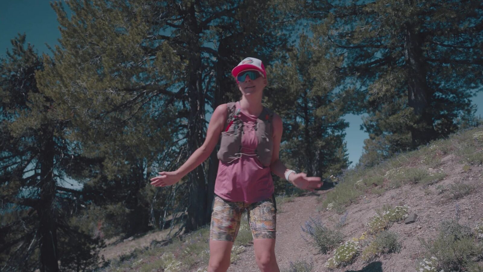 Joella Nicole standing on a desert mountain trail wearing sunglasses, a pink cap and a hydration vest